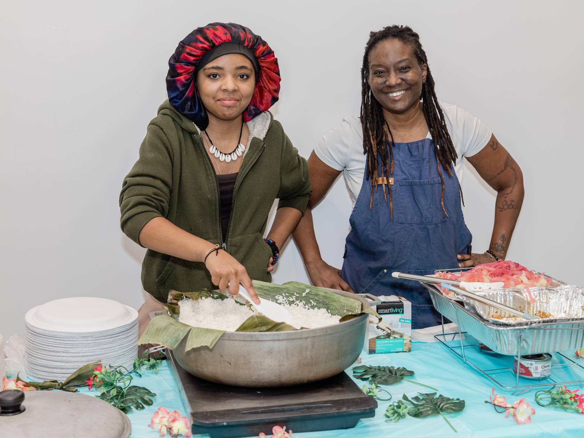 Two individuals stand next to each other standing behind a table full of different food items
