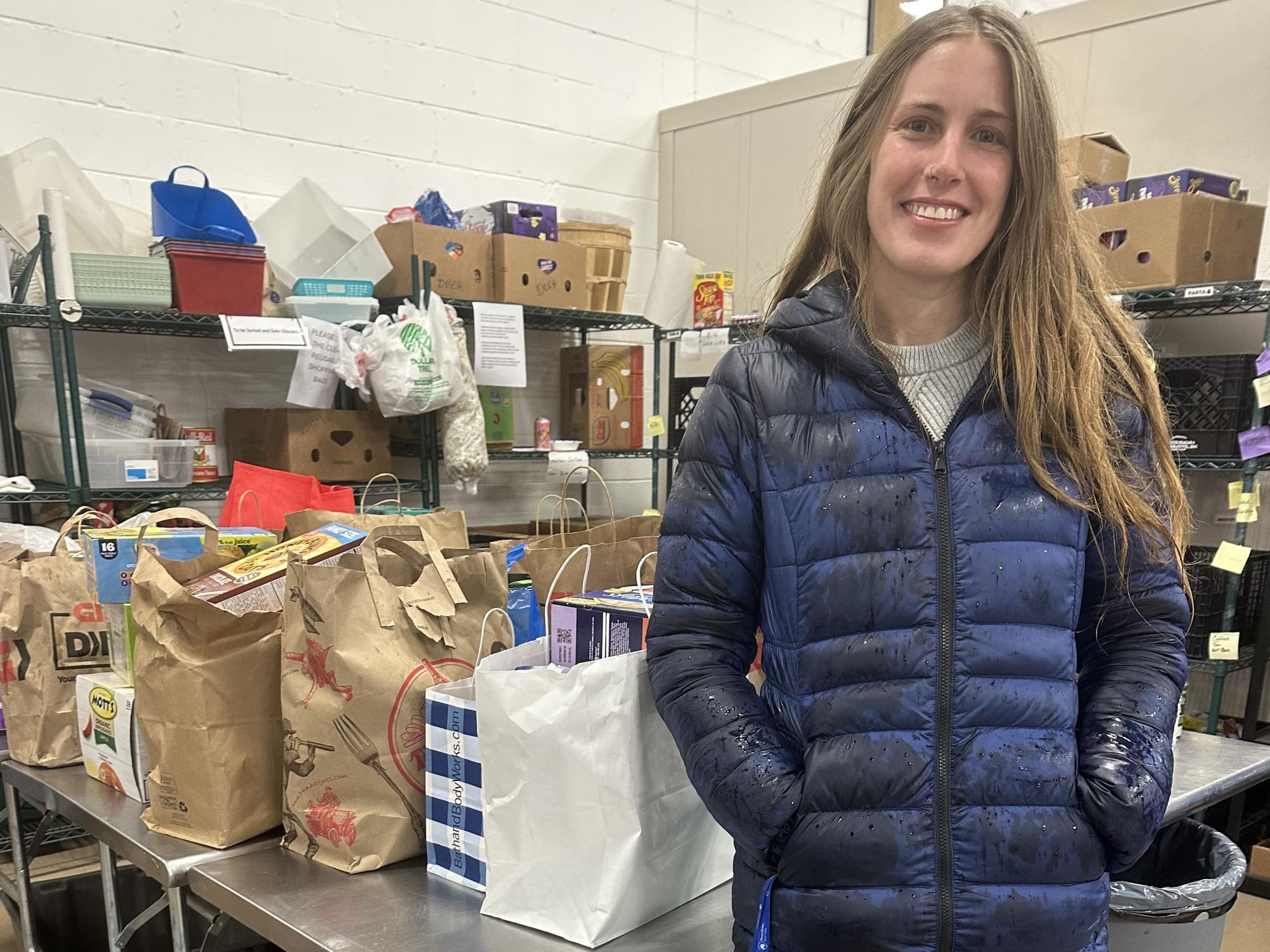 A smiling woman stands in front of a table and shelves with bags of donated food and personal care items.