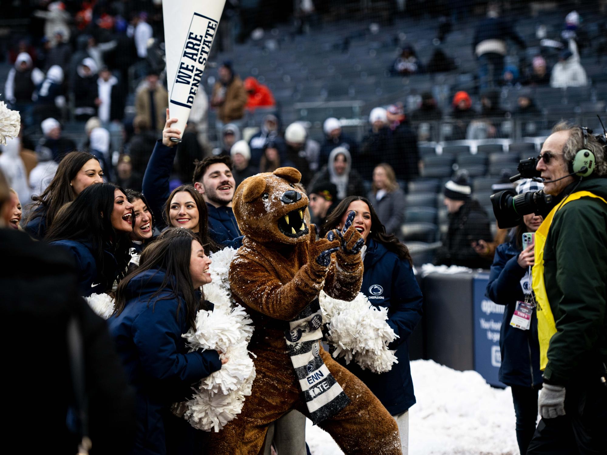 A group of cheerleaders and Nittany Lion Mascot pose and cheer in front of tv broadcast