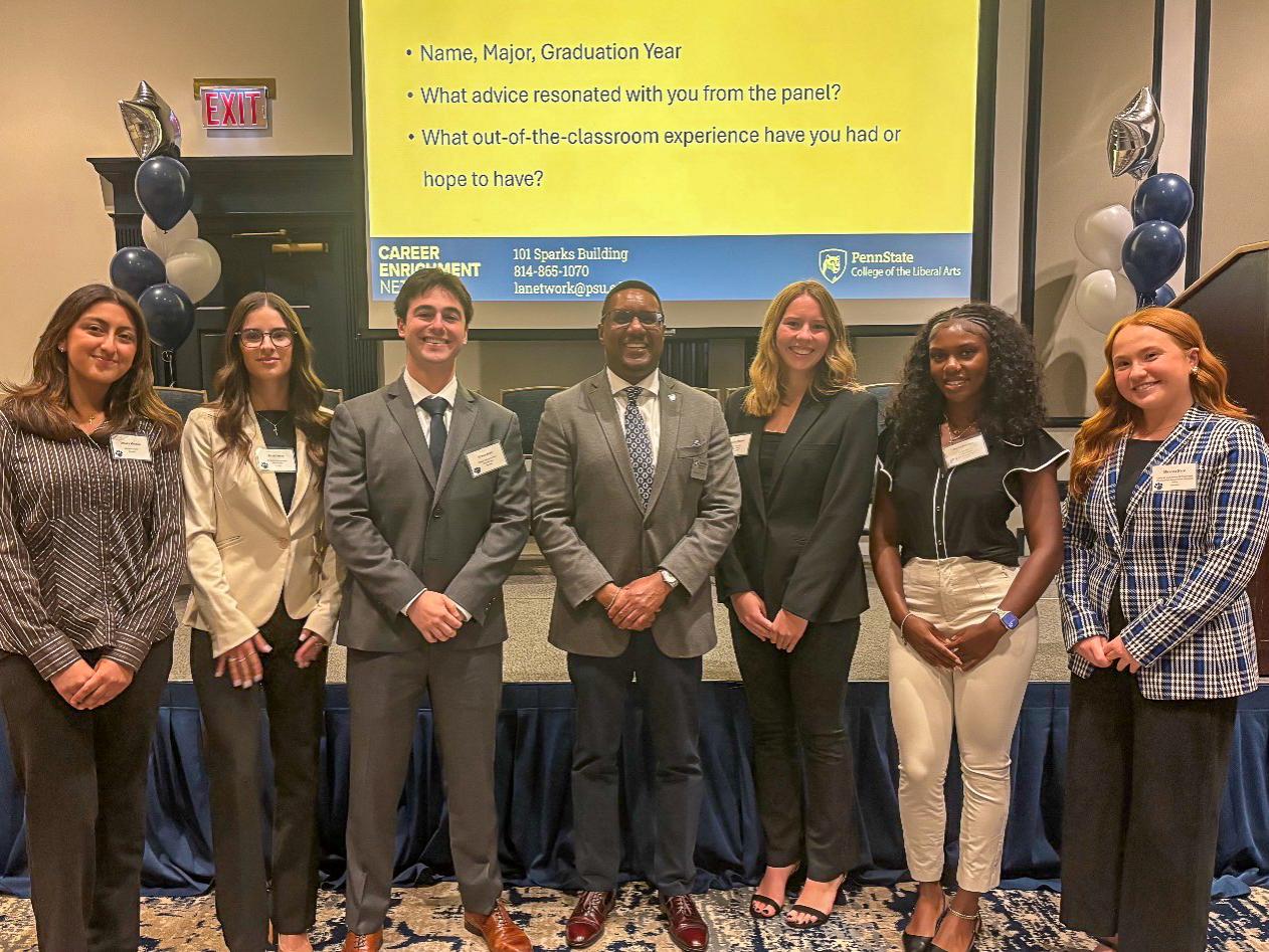 A group of students dressed in business attire posing together in front of a screen at a professional event or panel discussion.