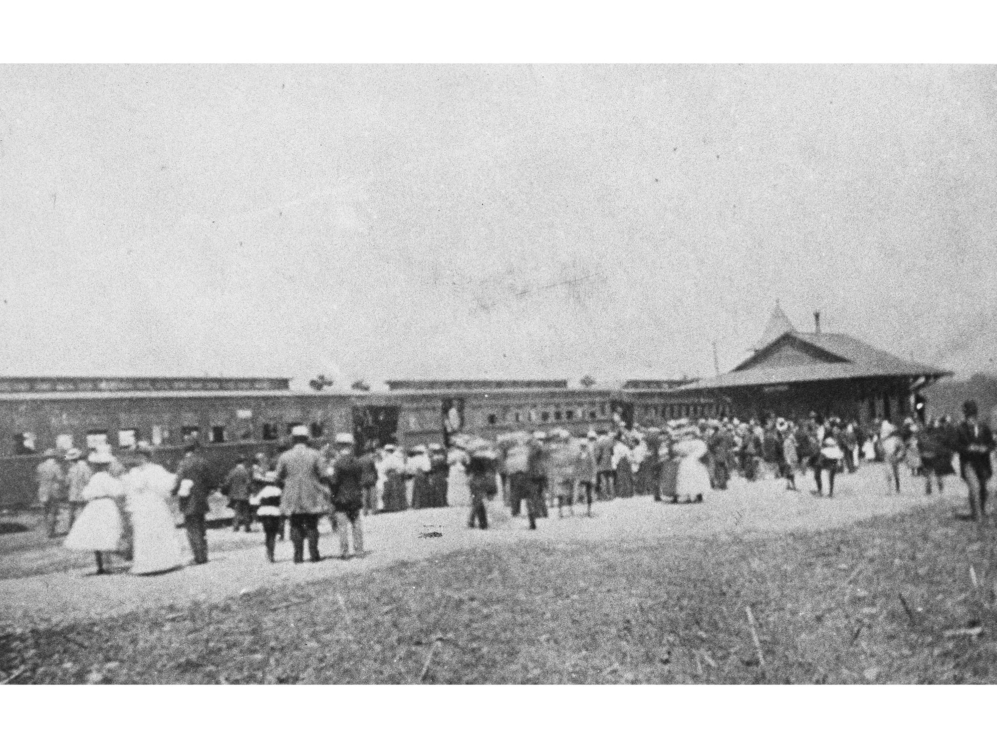 An historical image of a train at a station with people milling about