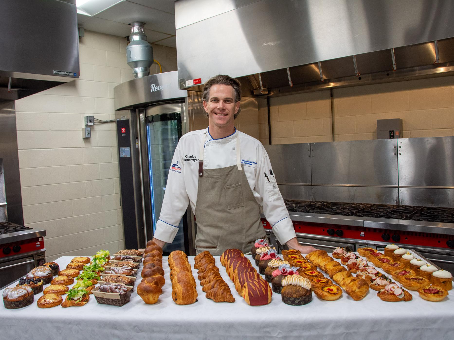 A smiling chef stands behind a table covered in delicious looking pastries