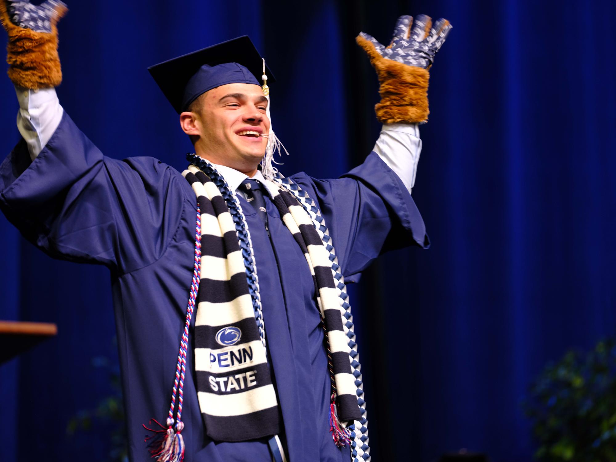 A smiling graduate wearing a navy cap and gown raises both arms in celebration on stage, wearing a Penn State scarf, honor cords, and fuzzy gloves.