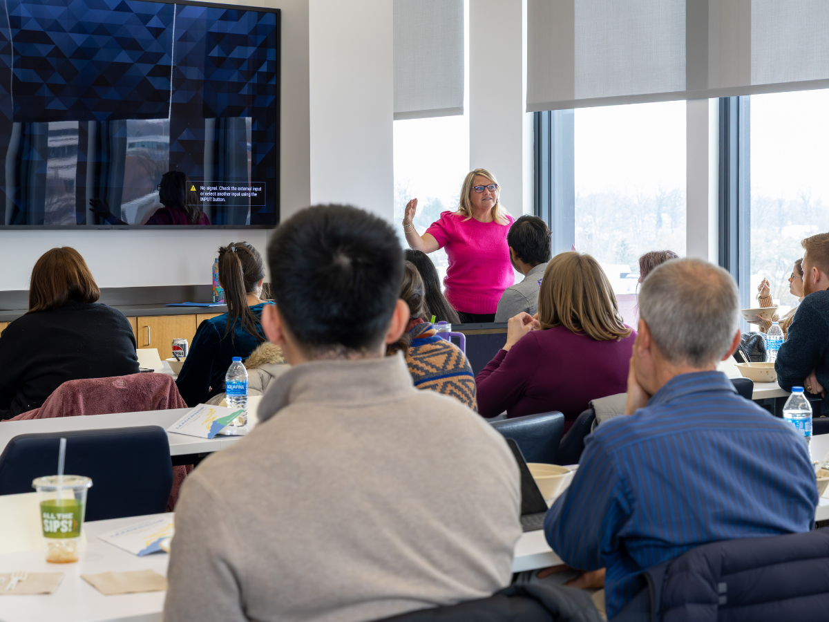 An individual standing in front of a room speaking to an audience of several seated individuals