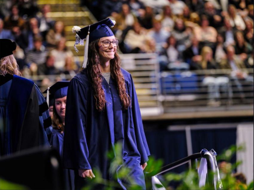 A student wearing a blue cap and gown walks across the commencement stage
