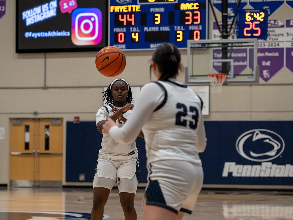 A Penn State Fayette women’s basketball player passes the ball to a teammate during a home game, with the scoreboard visible behind them.