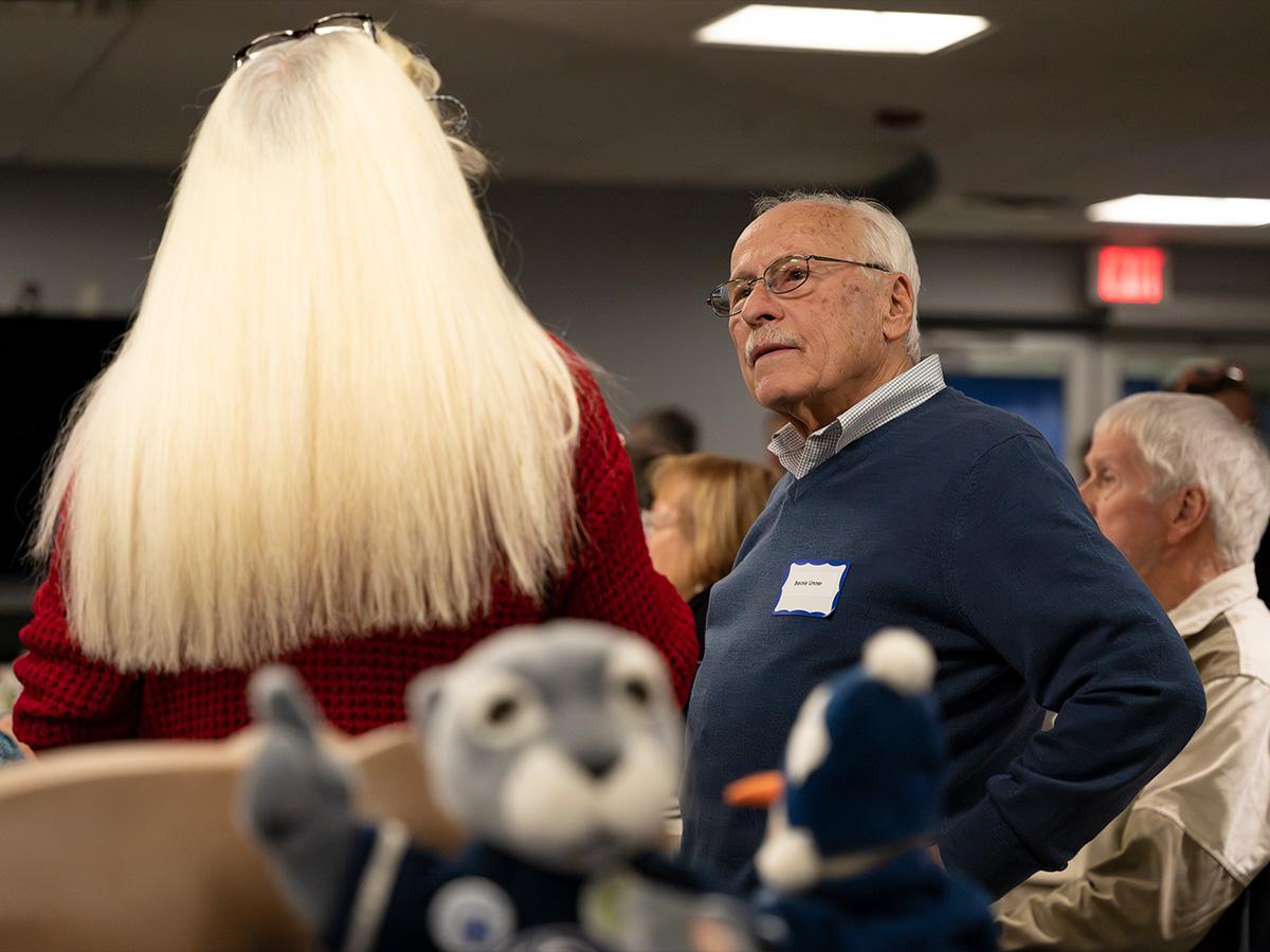 An older man wearing a name tag speaks with a woman in a red sweater during the anniversary event, with Nittany Lion plush toys in the foreground.