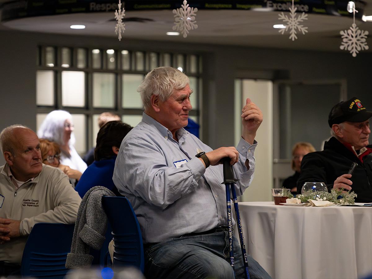 A man seated at a round table listens during Penn State Fayette’s 60th anniversary celebration, holding a cane as other guests sit nearby.