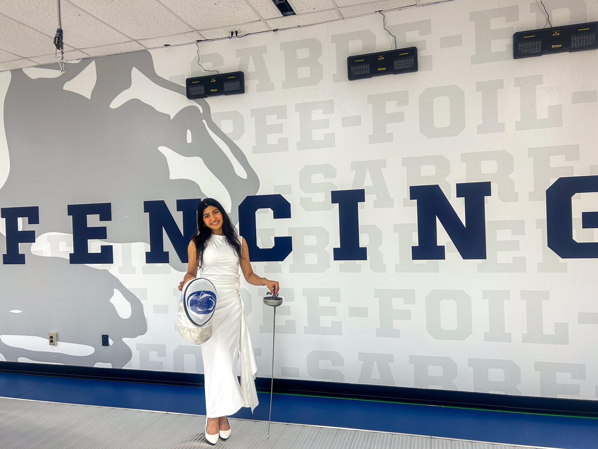 Vama Maniar standing in front of the Penn State Fencing wall in a white dress holding her gear.
