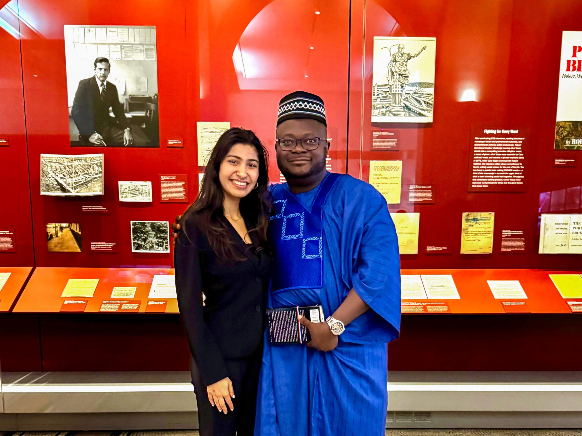 Vama Maniar wears a black suit and Samuel Kolawole wears blue traditional African clothing while standing in front of a display case. 