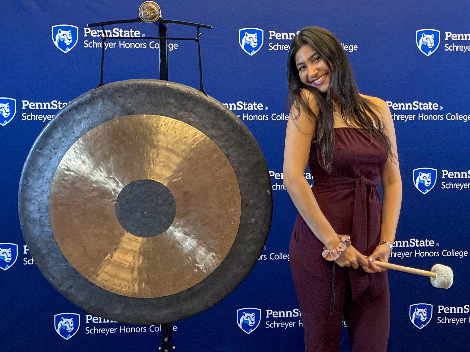 Vama Maniar stands next to the Schreyer Honors College gong in front of a navy backdrop with the Schreyer Honors College wordmark on it.