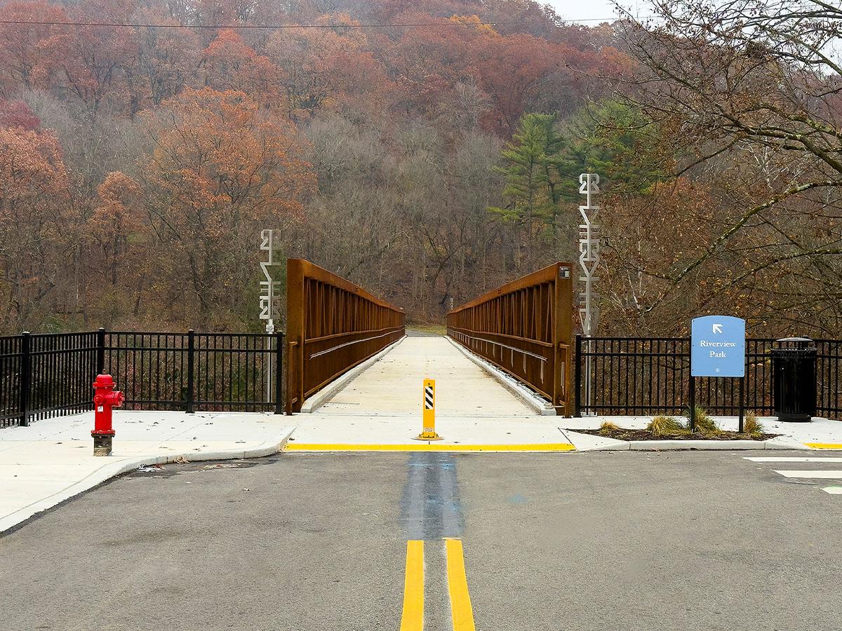 A pedestrian and bike bridge in Pittsburgh framed by rust-colored steel railings, with Hepner’s vertical metal sculptures installed at the entrance and wooded hillsides in the background.