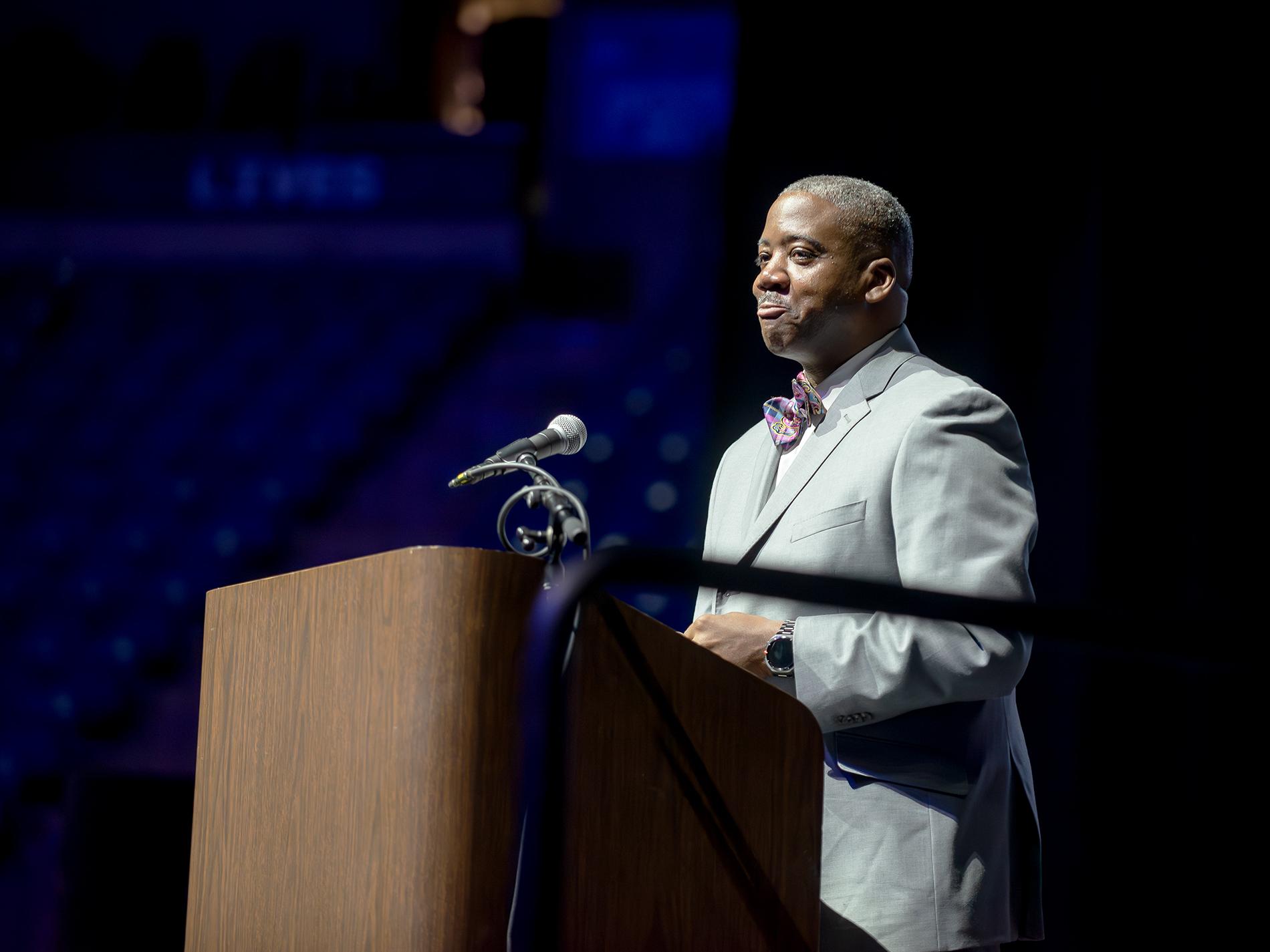 Jamie Campbell, wearing a light blue suit and colorful bow tie, smiles at the audience from the podium.