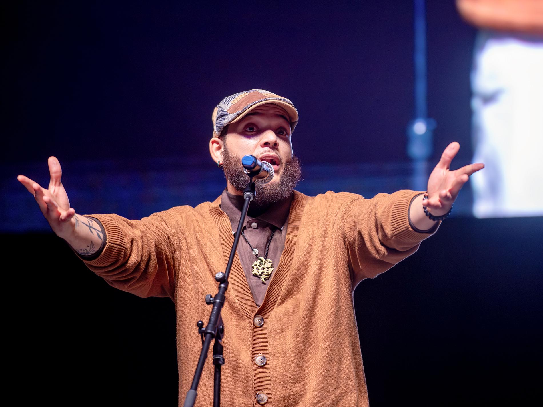 A person wearing an orange sweater and flat cap performs spoken-word poetry on a stage