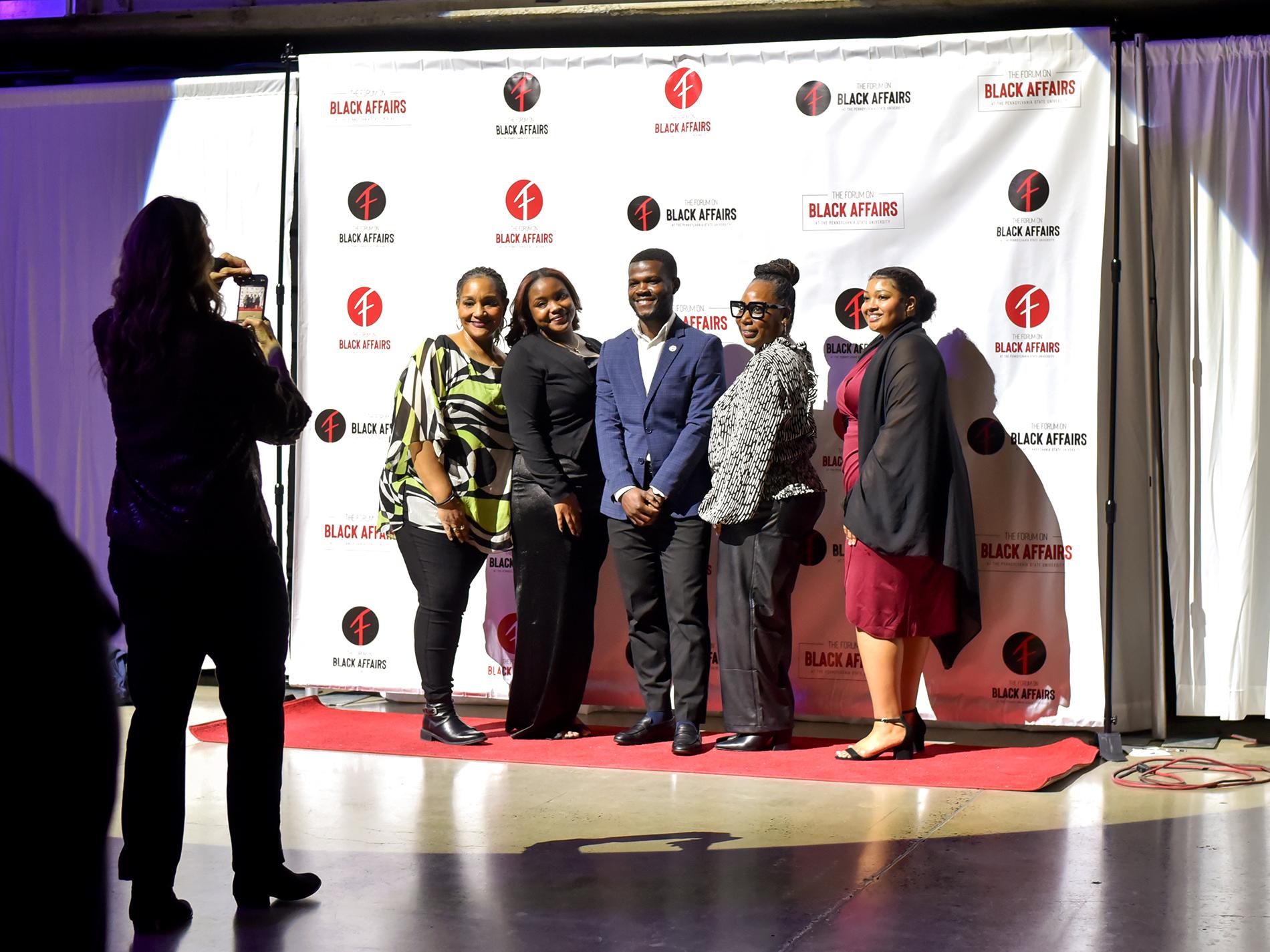 A group of five people pose for a photograph with a backdrop