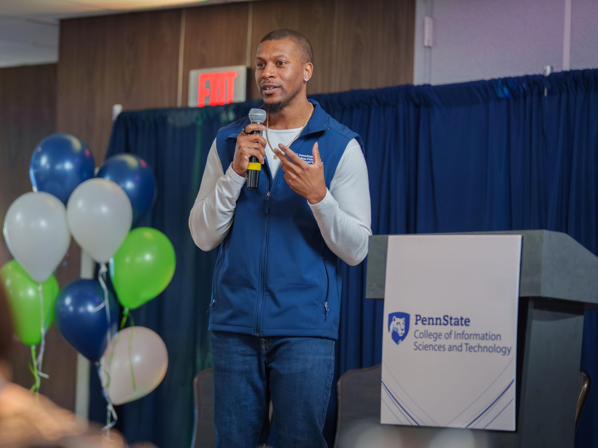 person wearing blue vest holding microphone, balloons and podium in background