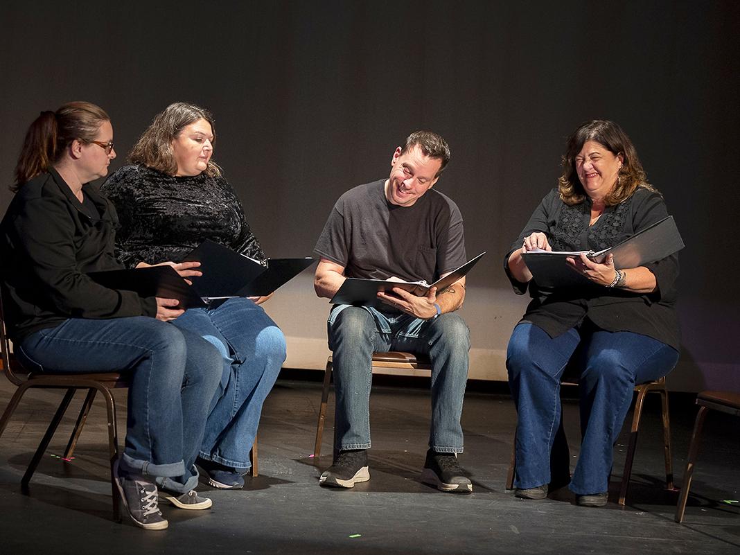 Four actors seated on stage reading from scripts during a rehearsal, reacting to dialogue under stage lighting.