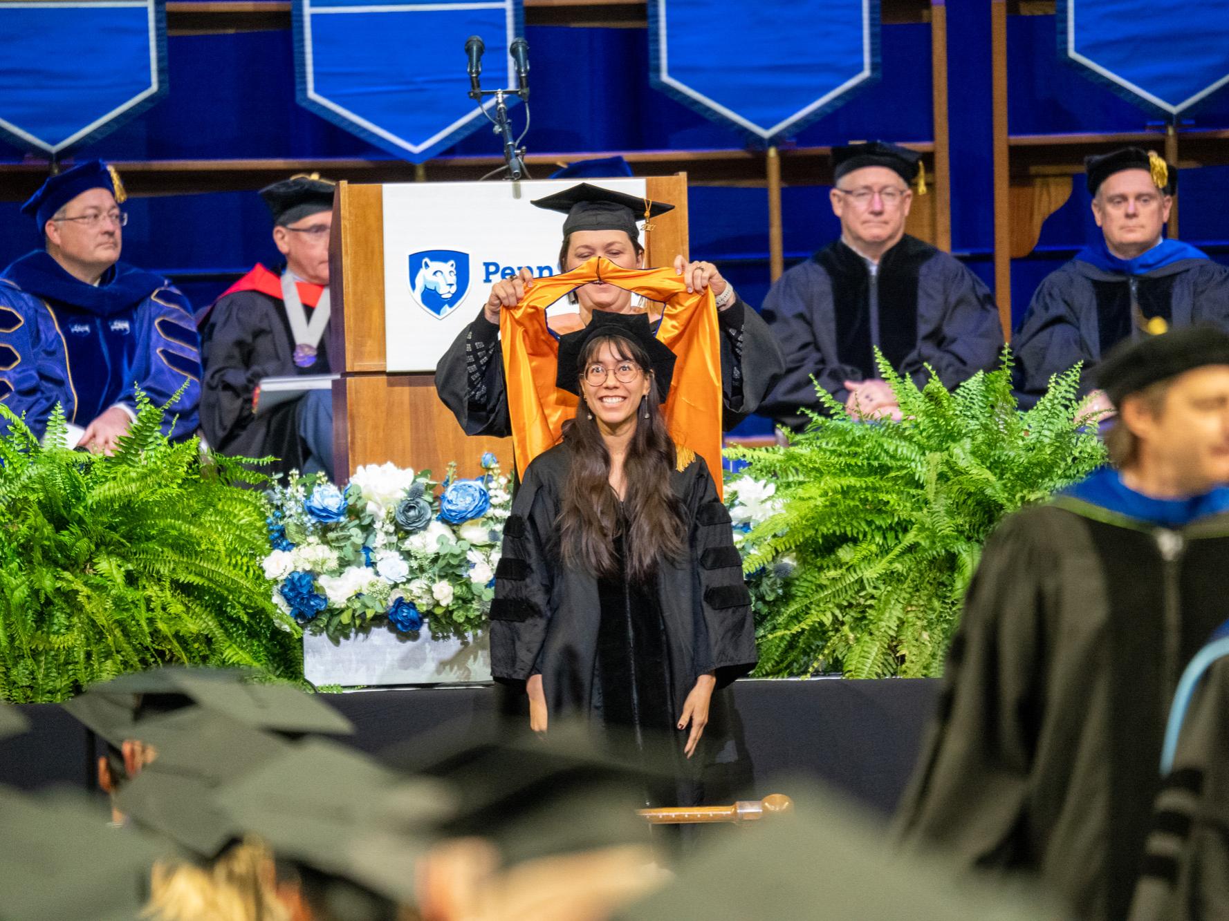 A woman stands on stage and waits as her commencement hood is placed over her neck.
