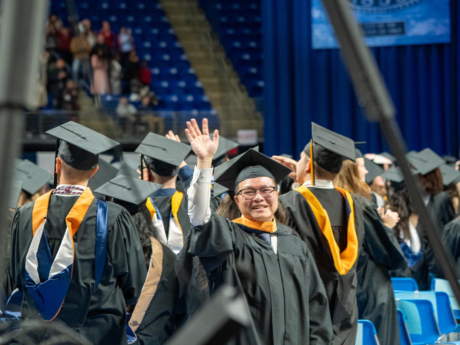 A man in commencement regalia waves toward the camera and smiles.