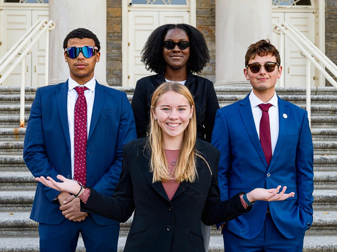 Shelby Corbett wears black and holds her arms out in front of a group of three people wearing sunglasses and standing in front of Penn State’s Old Main. 