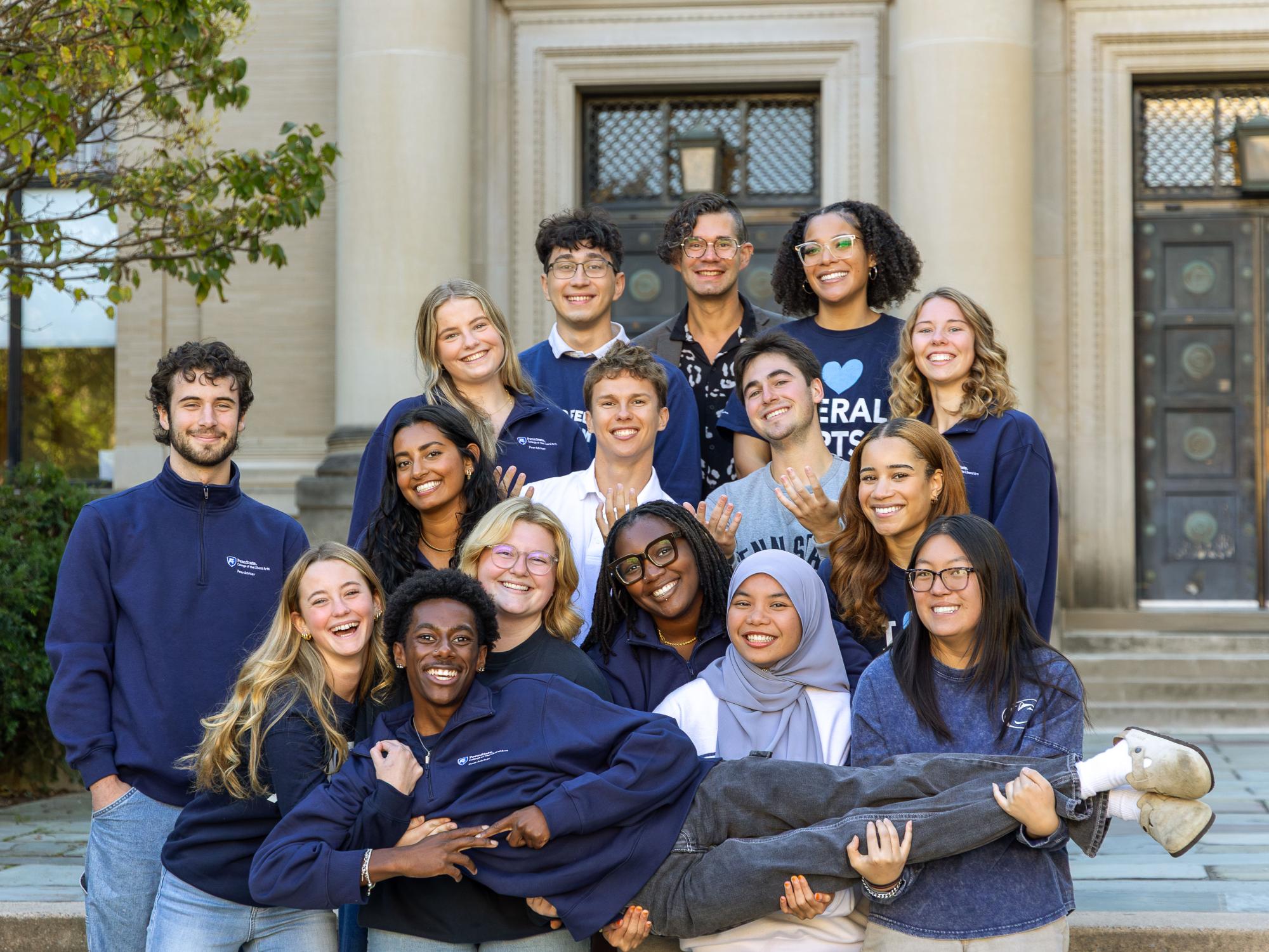 A group of peer advisers wearing navy sweatshirts stand in front of Sparks Building. They hold one of the advisers who is "laying".