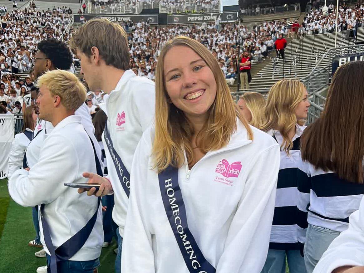 Shelby Corbett wears a white quarter-zip sweatshirt and a Homecoming Court sash while standing on the field at West Shore Home Field at Beaver Stadium.