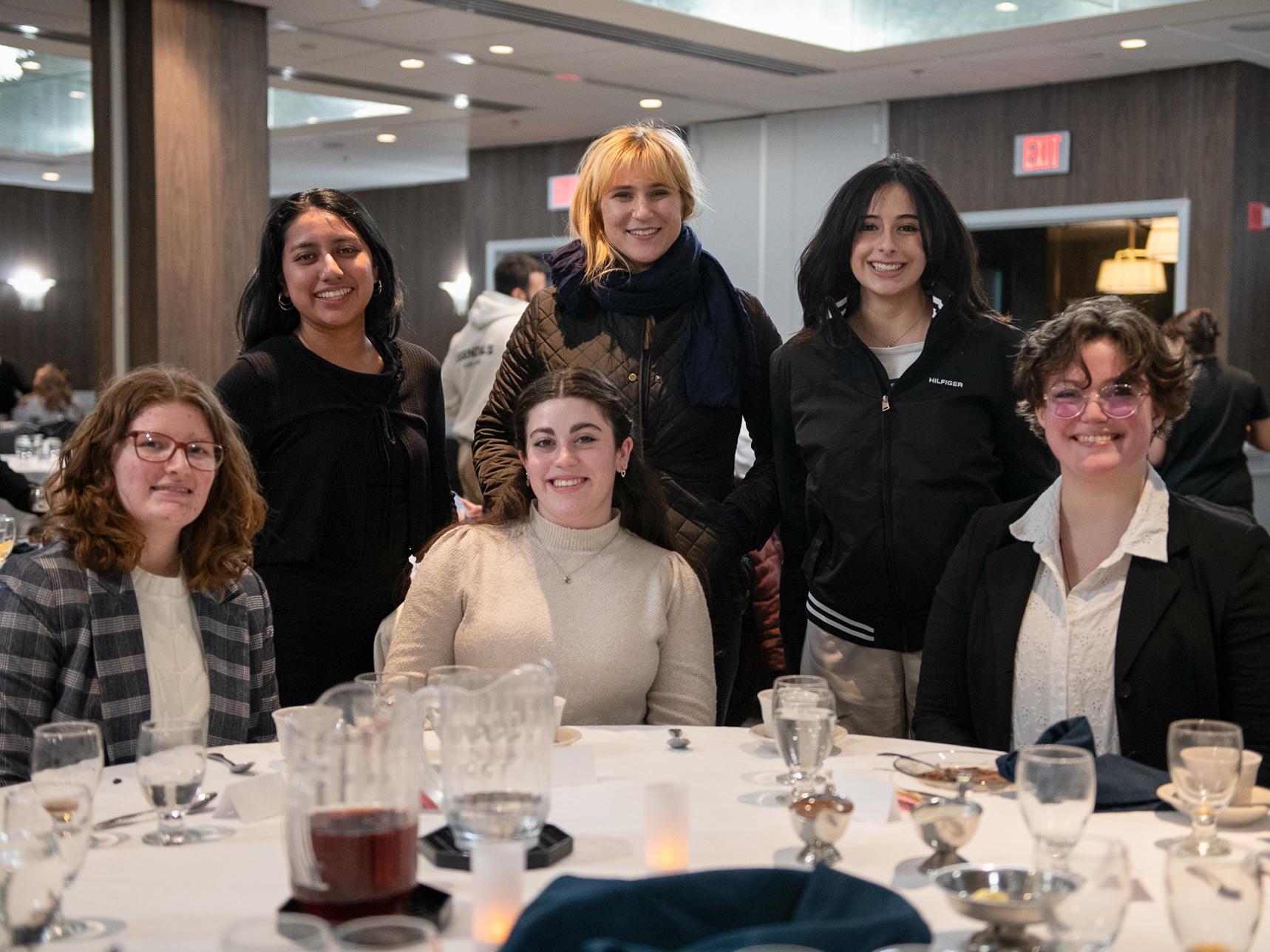Six Penn State students sit and stand around a dinner table at the Etiquette Dinner during Love Liberal Arts Week in February 2025.