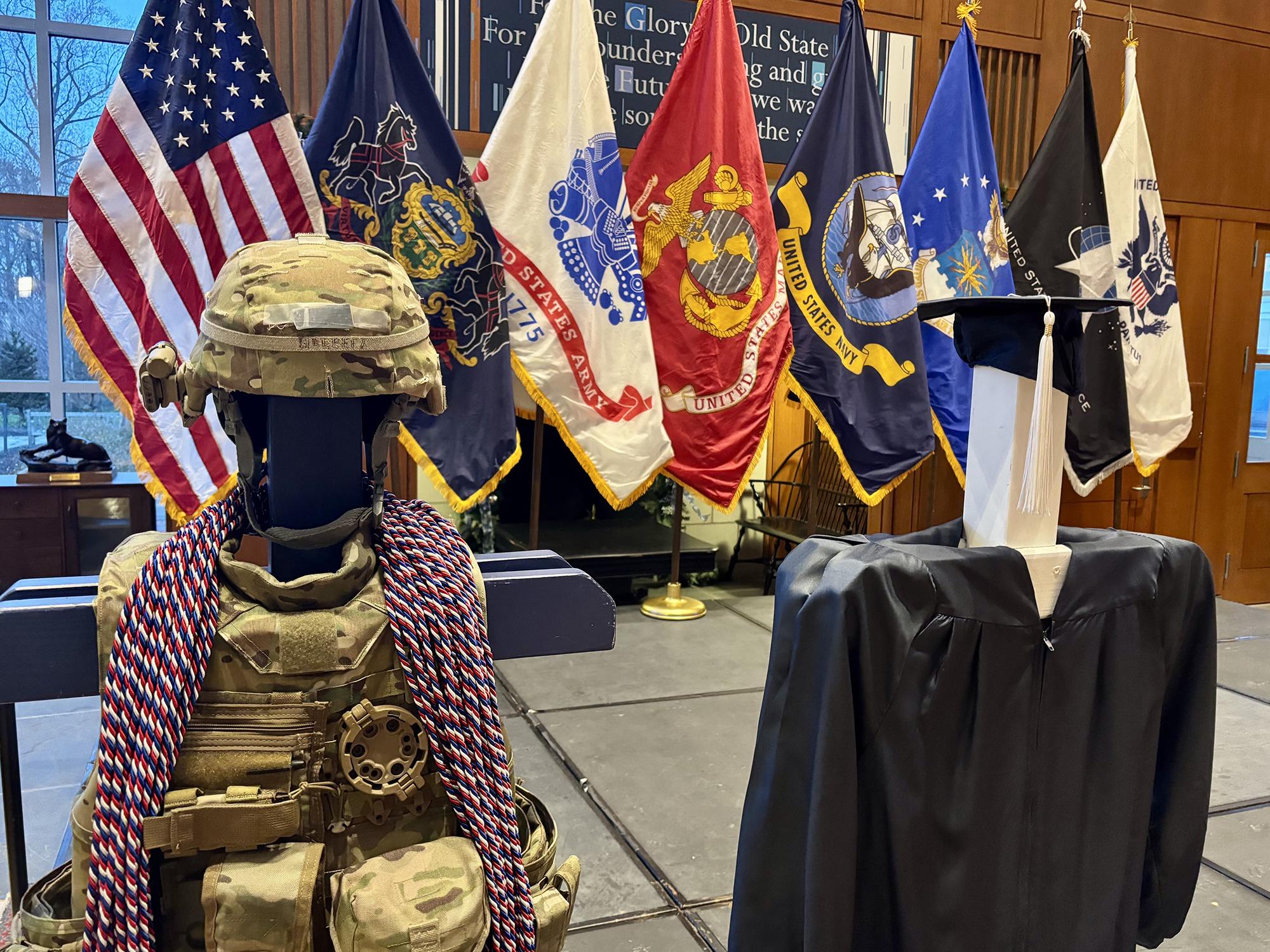 Red, white, and blue honor cords lay on the shoulder of camouflaged military combat gear in front of U.S. military flags.
