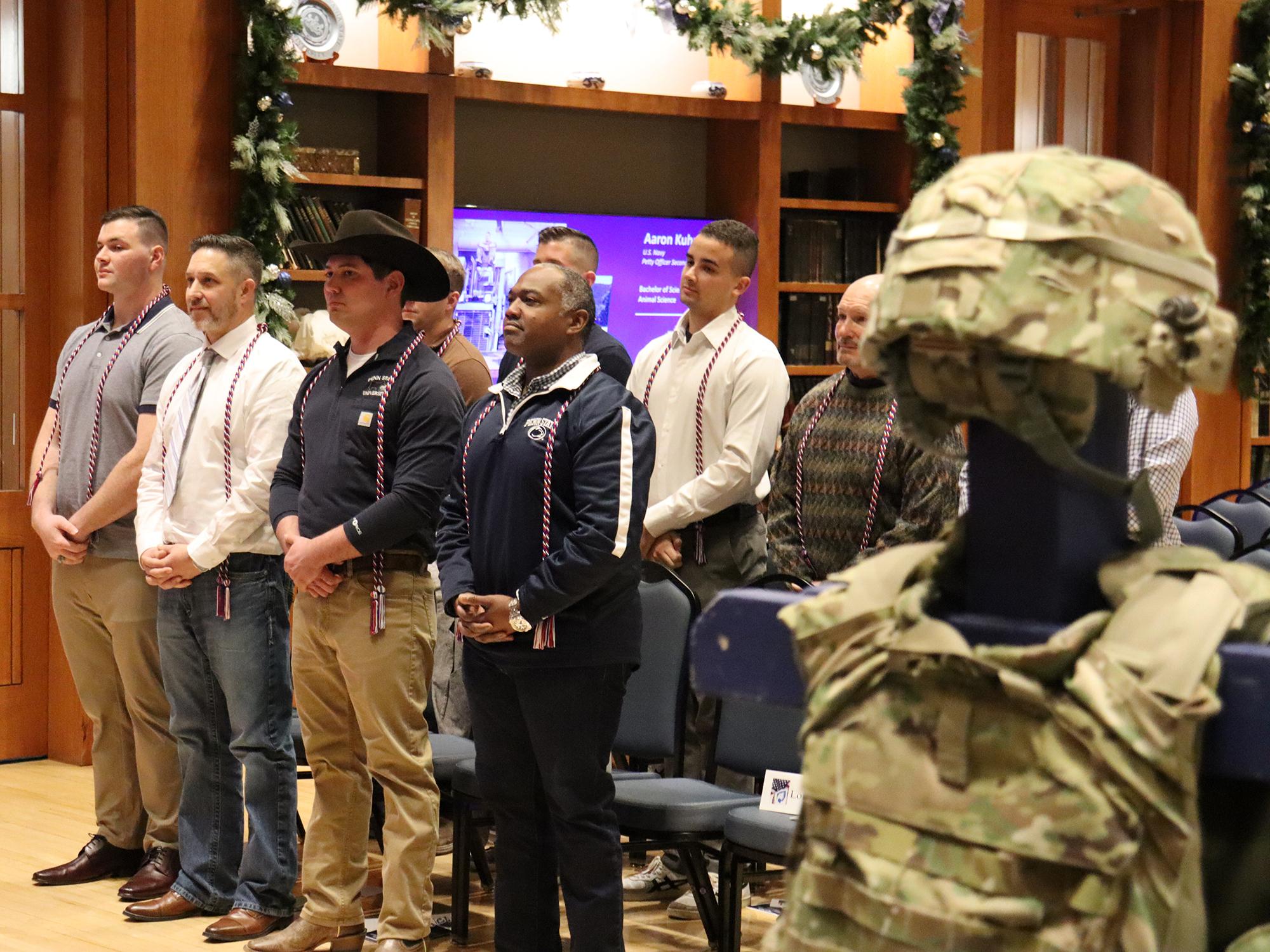 Camouflaged military body armor is displayed in a room full of people wearing red, white, and blue honor cords.