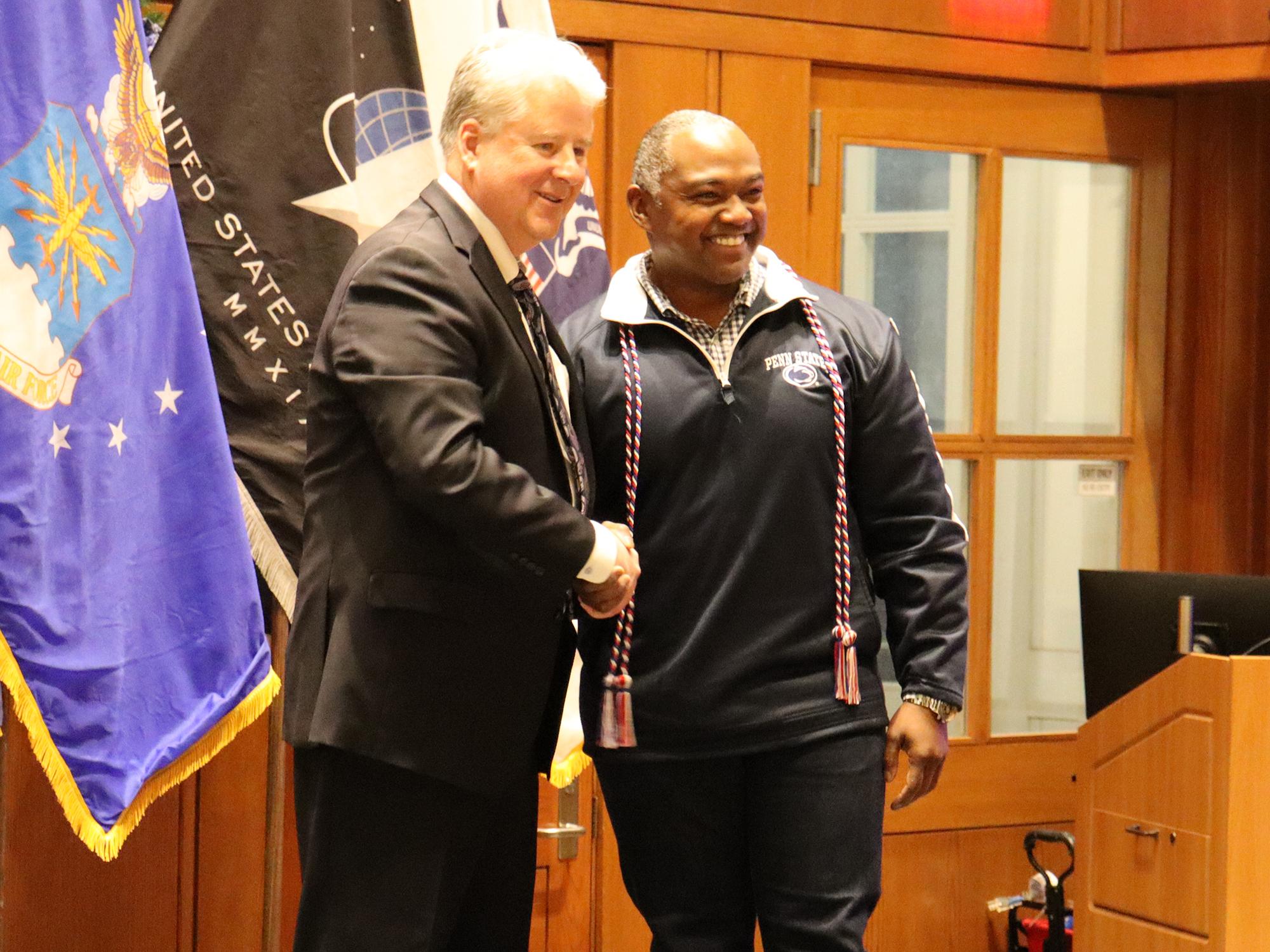 Two men shake hands in front of U.S. military flags.