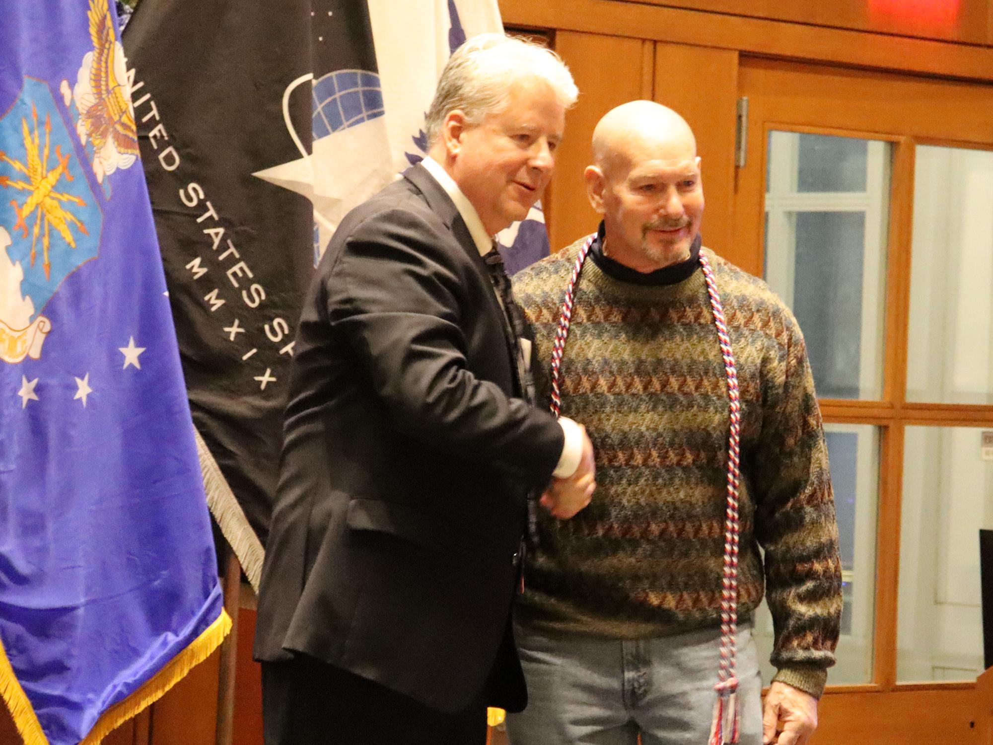 Two men shake hands in front of U.S. military flags.