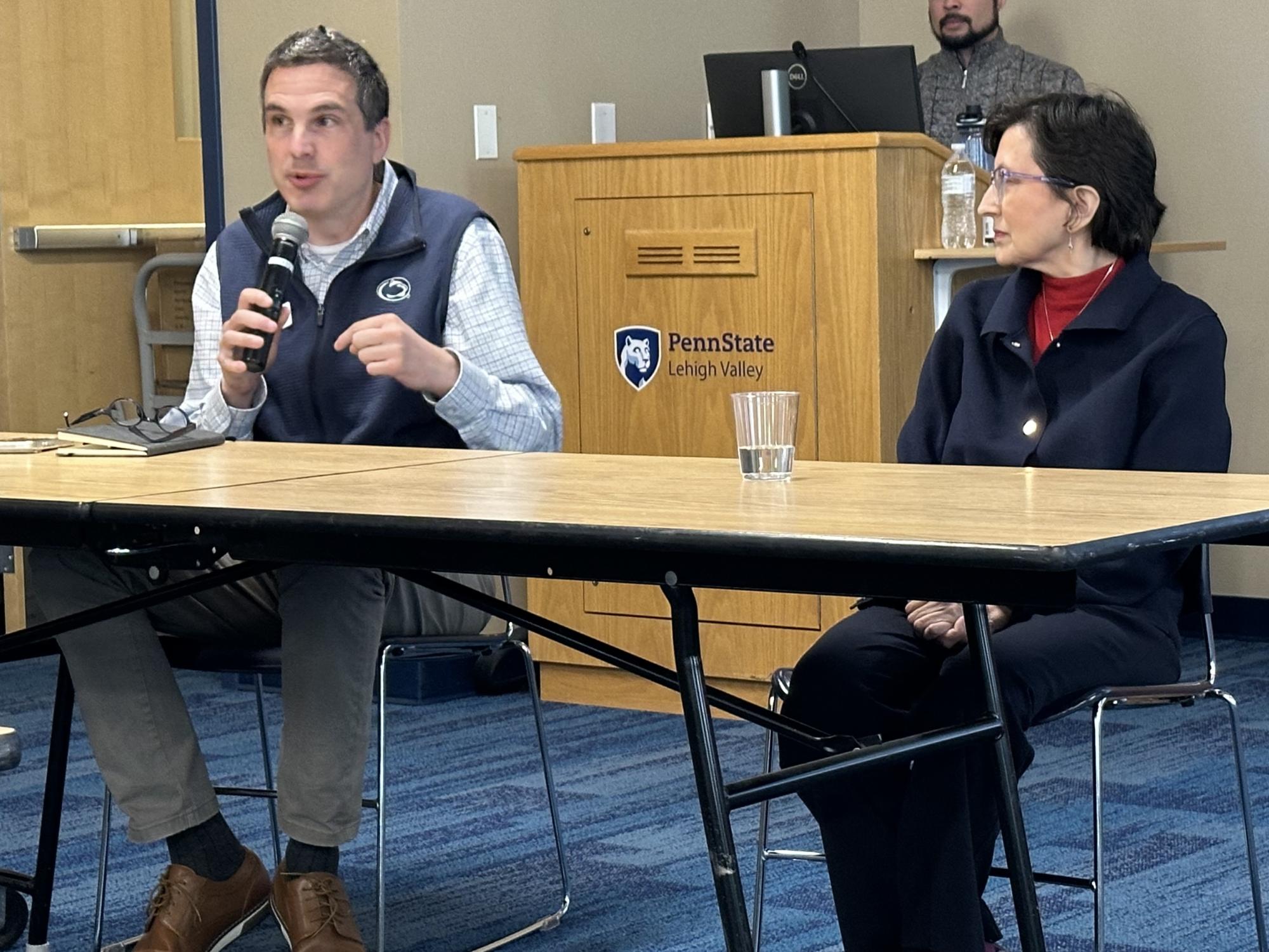 two people sitting at a table during a panel discussion, one is speaking into a microphone