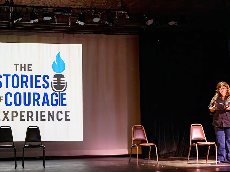 Person reads from a script on stage beside empty chairs, with a projected sign reading The Stories of Courage Experience.
