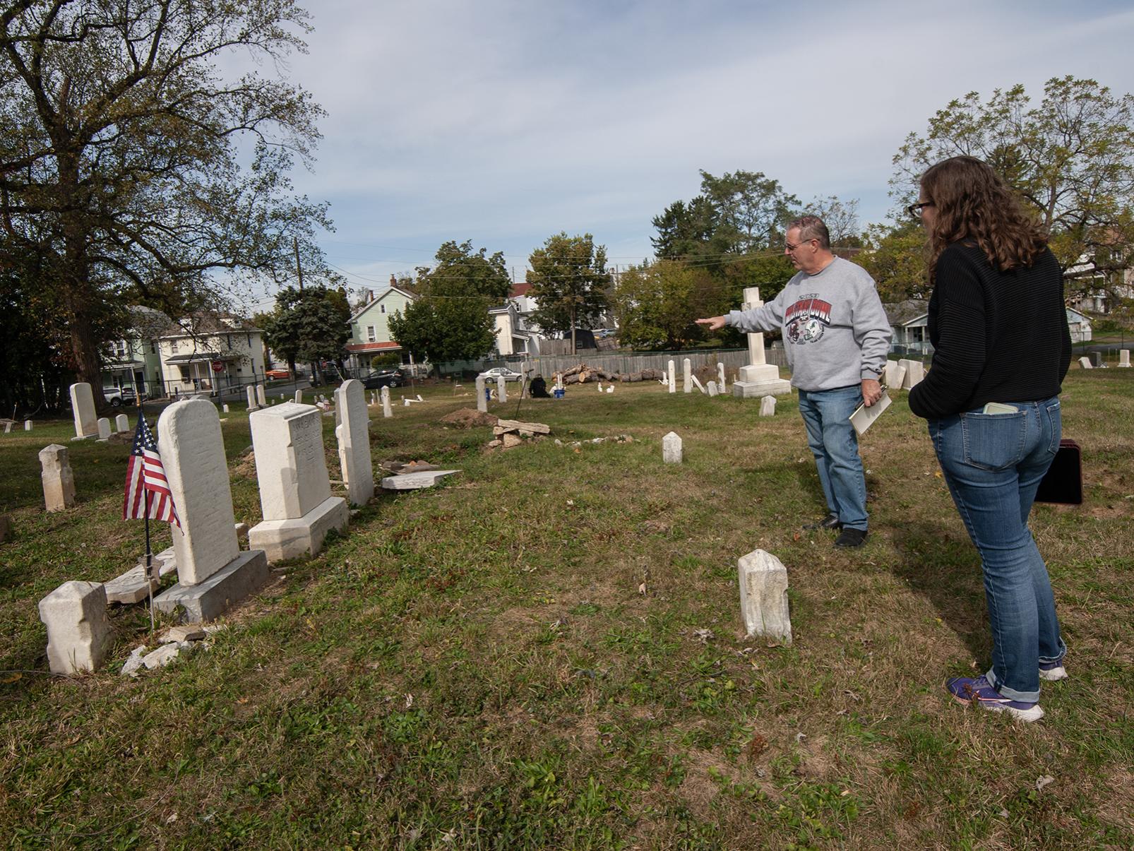 A man points to a headstone in a cemetery, while a woman looks on