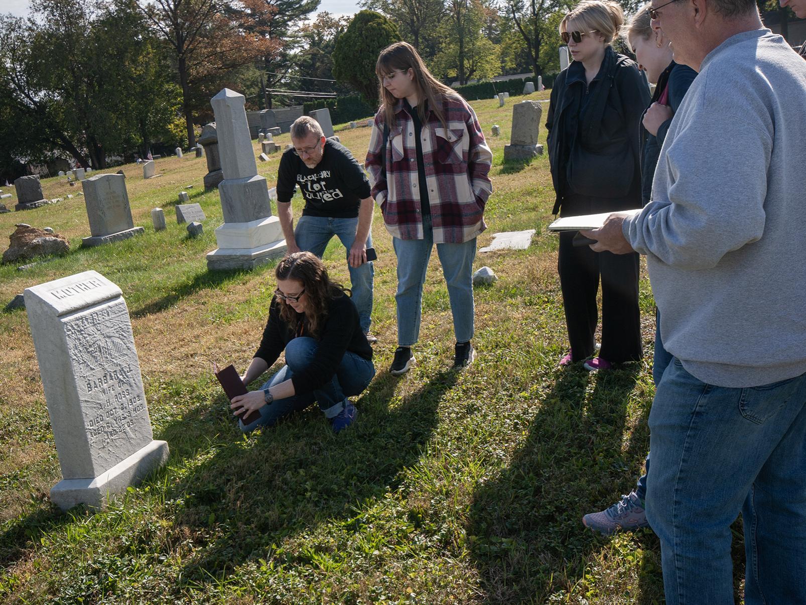 A woman uses a device to 3D scan a headstone in a cemetery, while a group of college students gather to watch