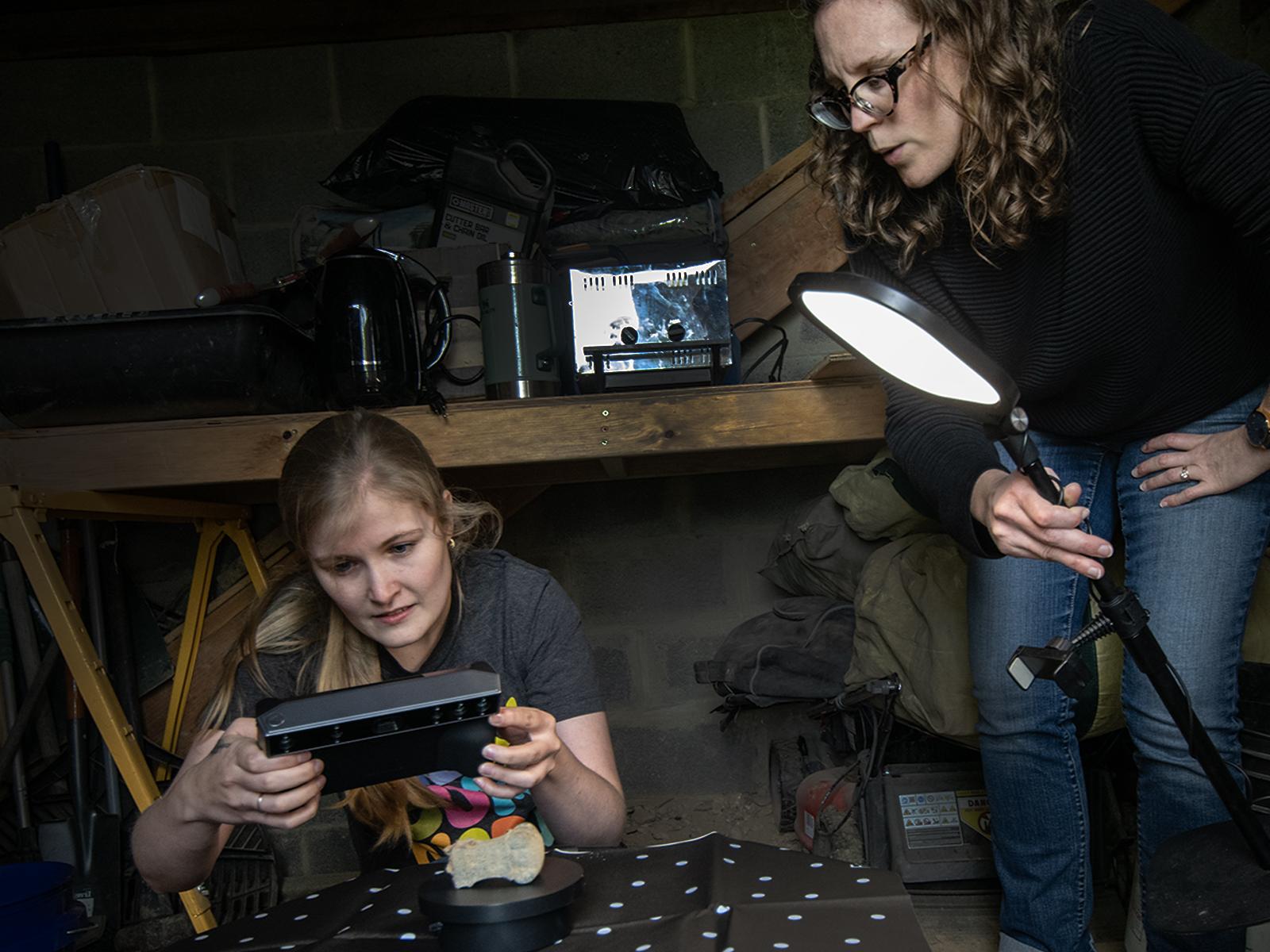 A woman uses a portable 3D scanner on an object, while another woman holds a light over the object