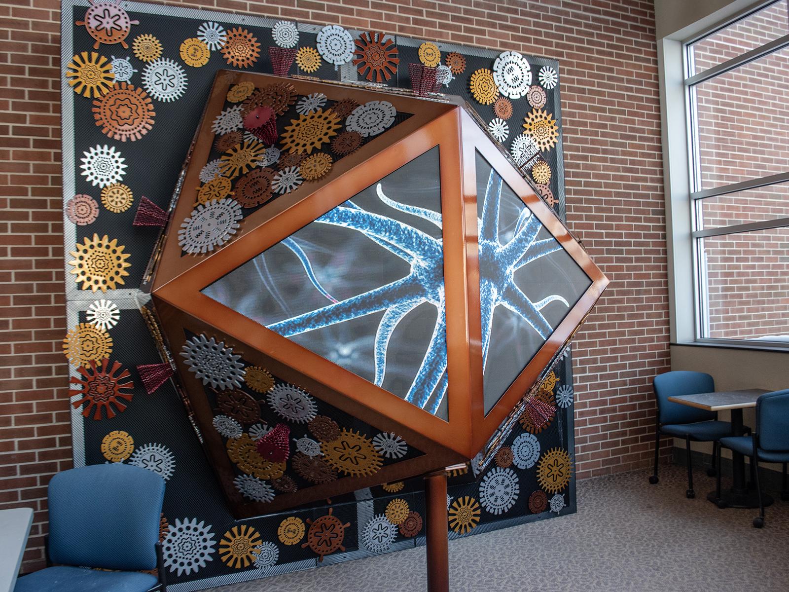 A large art installation featuring aluminum gear-like shapes and LED screens showing videos and visualizations related to virus research