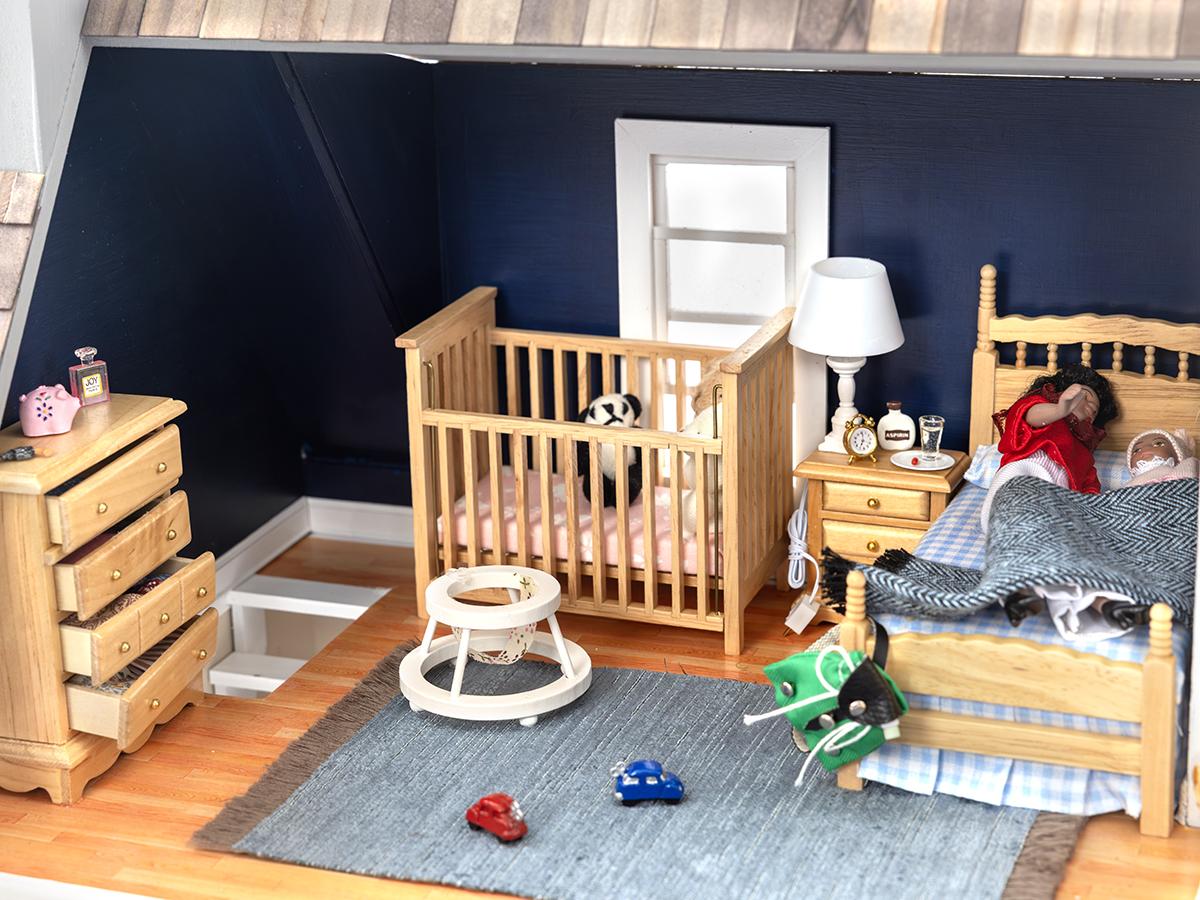 A dollhouse bedroom shows a mother sleeping with a baby, a walker on the floor and a stuffed animal in a crib.