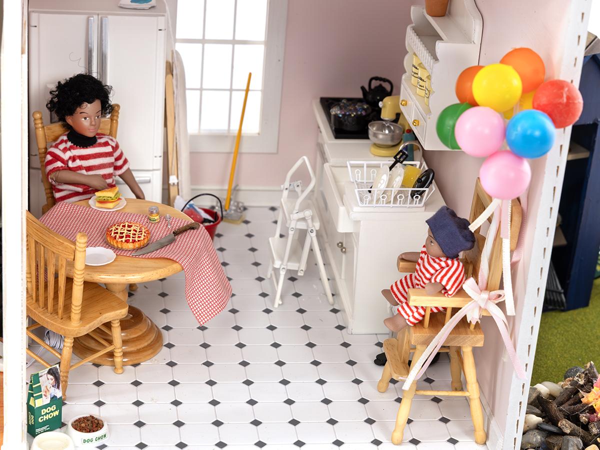 A dollhouse kitchen shows a man sitting at the table and a baby unsecured in a high chair.