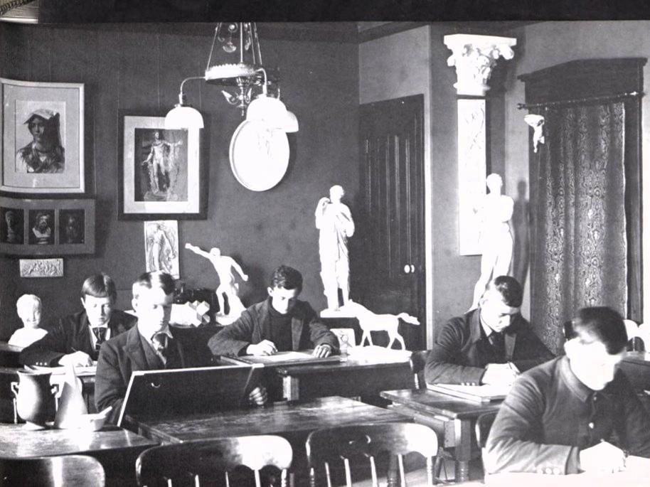 A black and white photo of students at desks drawing on paper in the 1890s