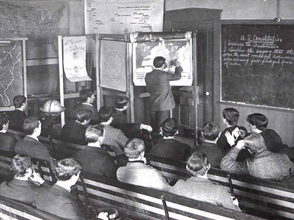 A black and white rear shot of students in a classroom in 1894 while an instructor points to a map