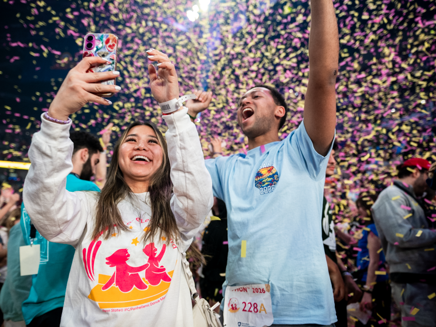 Two Penn State THON participants celebrate under falling pink and gold confetti in a crowded arena, smiling and cheering as one person holds up a phone to take a selfie.