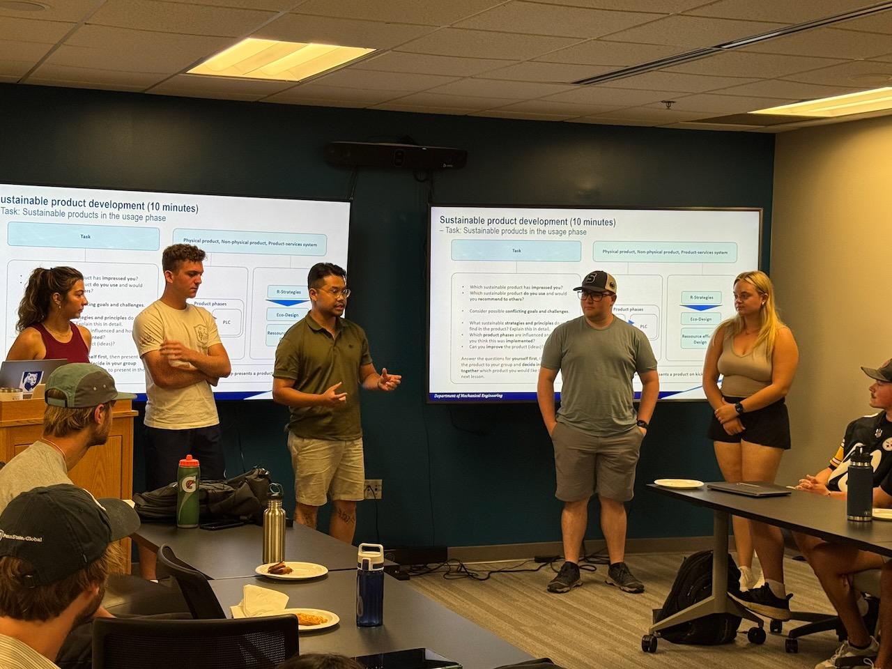 Five people giving a presentation in front of TV screens
