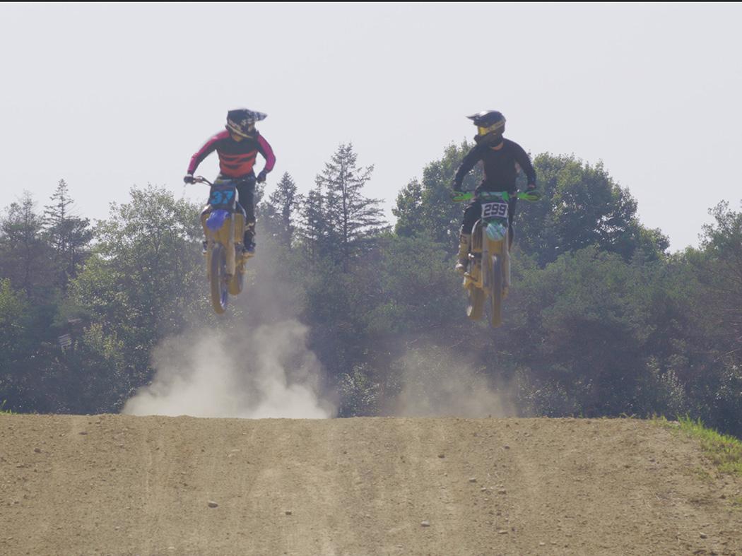Two motocross cycles take to the air over a small mound of dirt