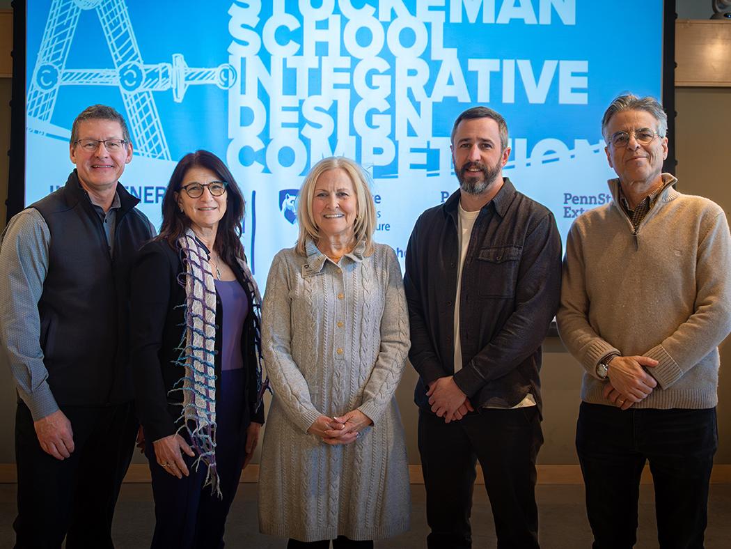 Five people stand together in front of a large blue event screen displaying the text “Stuckeman School Integrative Design Competition.” They are posed in a row, dressed in business‑casual attire, with the event graphics and logos visible behind them.