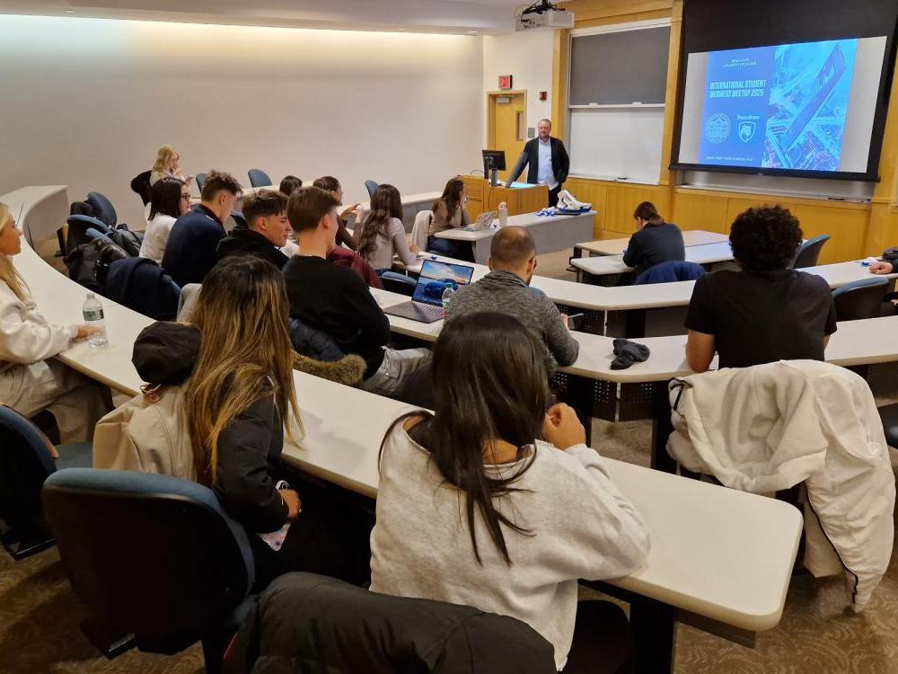 A speaker talks to students sitting in a classroom and watching over a virtual meeting.