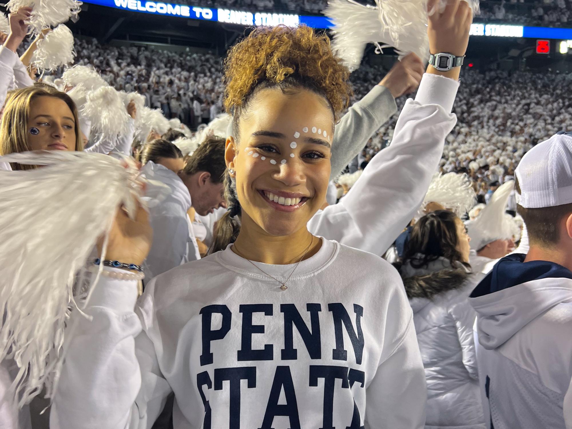 Alivia Peterson wears a white sweatshirt that reads “Penn State” while waving a white pom pom and smiling at the camera during the Penn State White Out football game in 2022.
