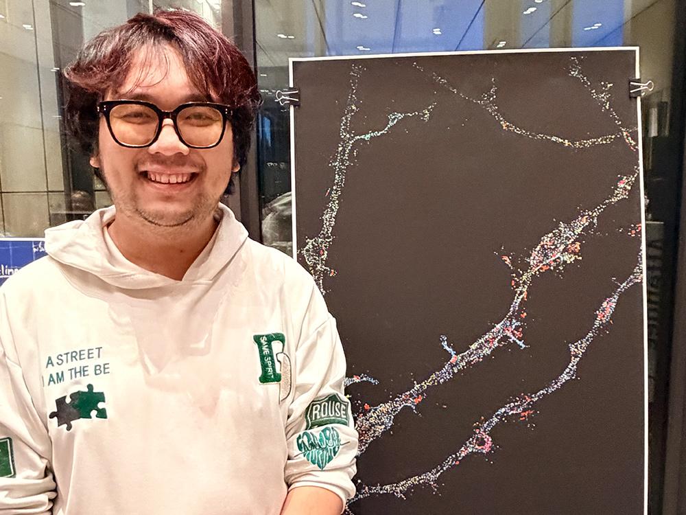 A photo of Yuanmin Zheng standing beside a print of his artwork, which is primarily black, with a series of thin, colorful, branching lines.