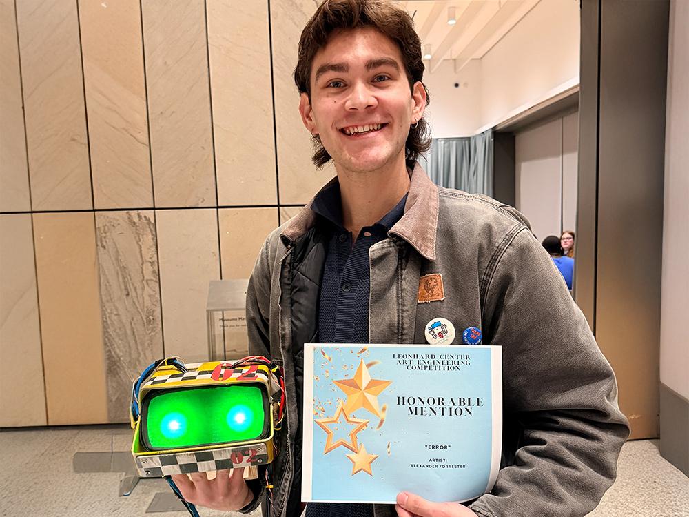 Alexander Forrester holding an honorable mention award in one hand and his artwork in the other—a small mechanical object that looks like a virtual reality headset, with two green glowing lights emanating from a screen and electrical wires running down either side of the machine. 