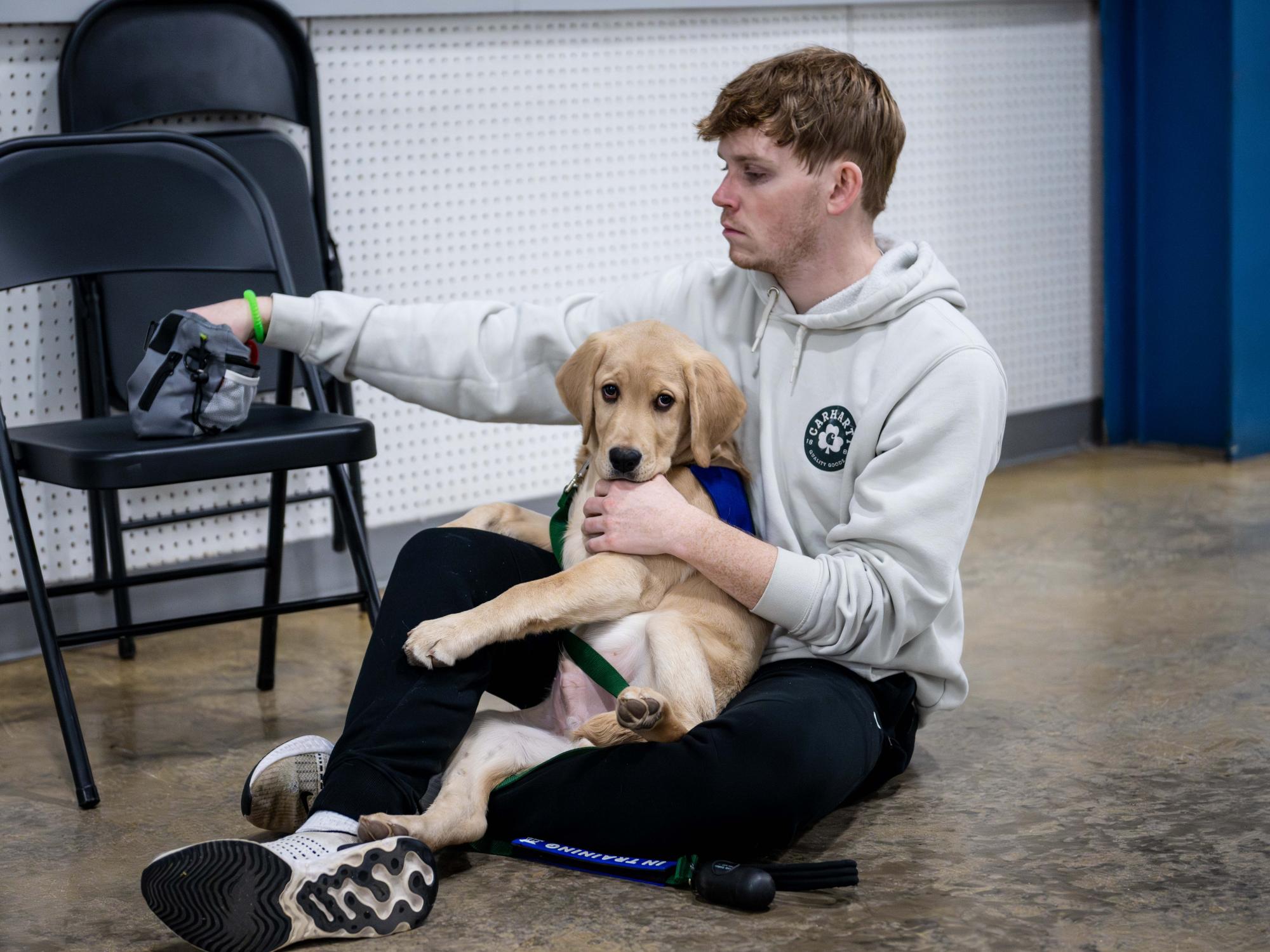 A student sits on the floor holding a golden retriever puppy wearing a purple training vest during a service dog training session.